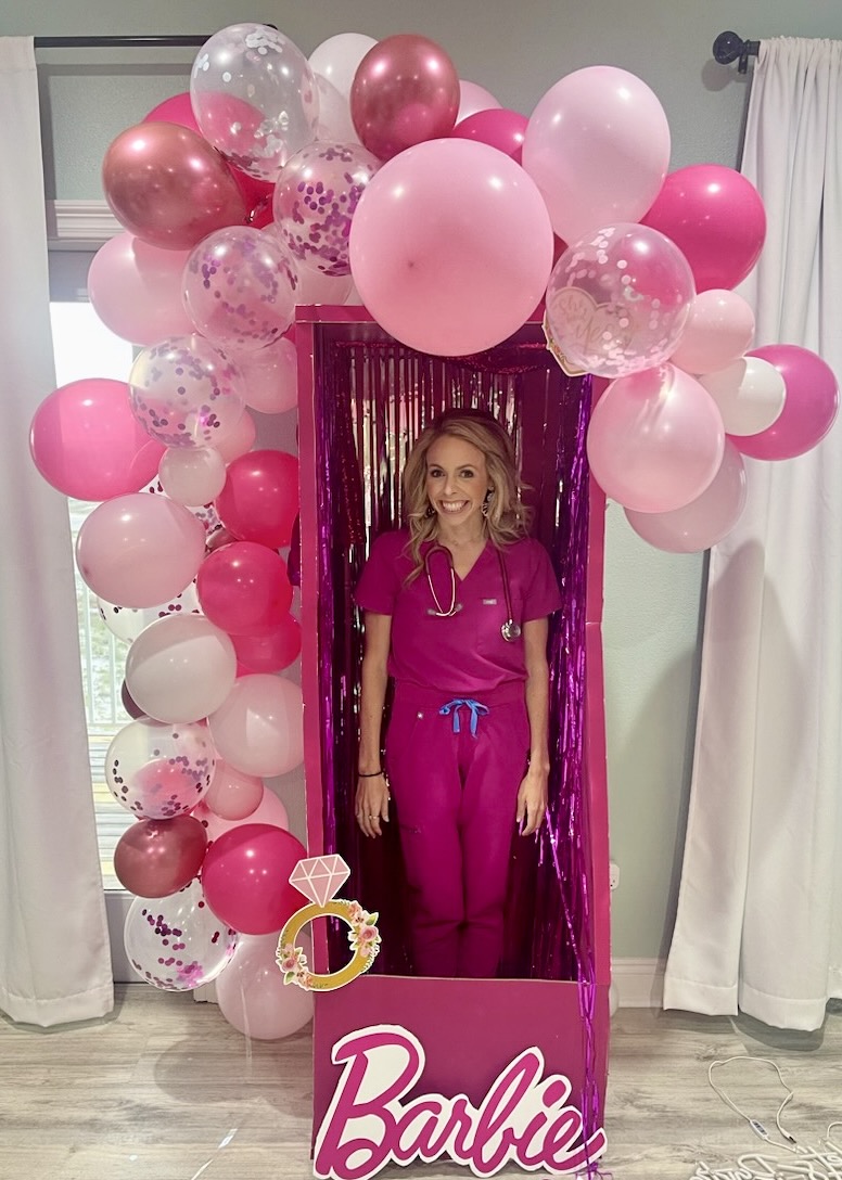 Dr. Carpenter wears hot pink scrubs while standing inside a &quot;barbie&quot; box draped with pink balloons