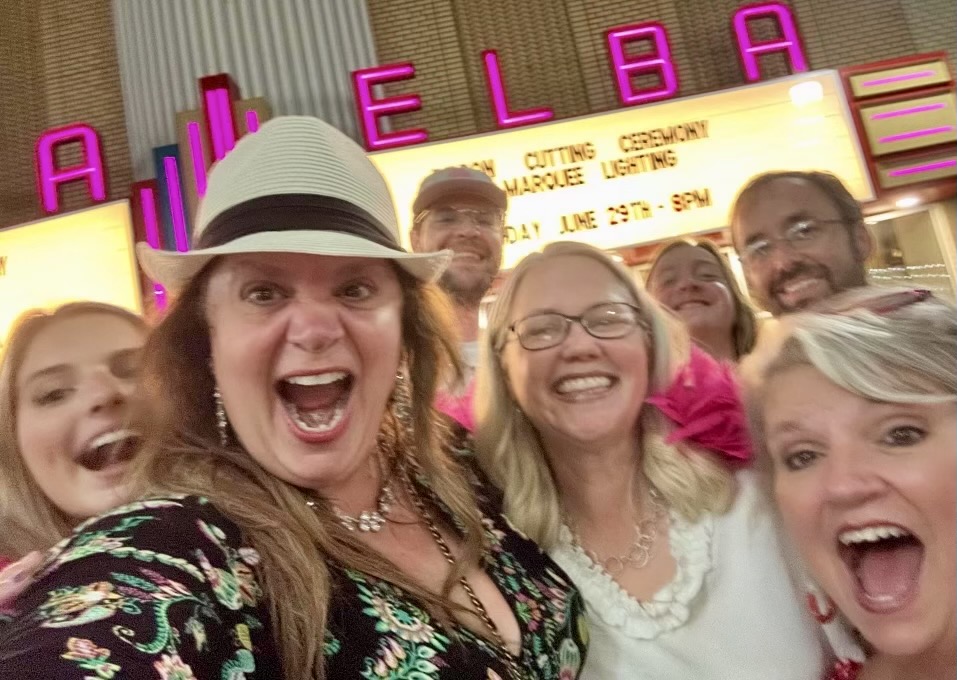 Marilyn Chivetta wears a fedora while posing with friends in front of the illuminated Elba Theatre marquee