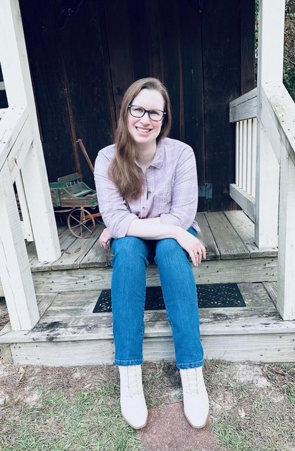 A young woman with long brown hair and glasses sits on the wooden steps of a rustic porch, smiling at the camera.