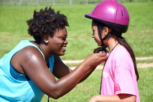 a young woman in a blue shirt fastens the helmet of a girl in a pink shirt in preparation to ride