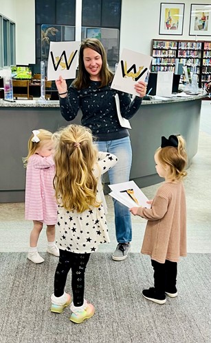 A library volunteer holds up two pages with the letter "W" for three young children