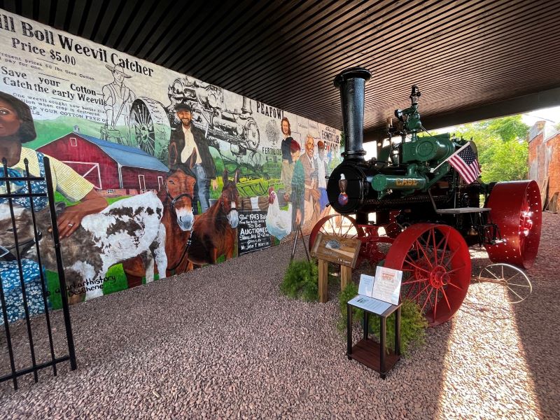 Steam tractor standing in pavilion where agricultural collage mural is displayed on wall